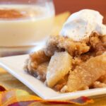 A plate of apple crisp with a dollop of whipped cream and some eggnog in the background.
