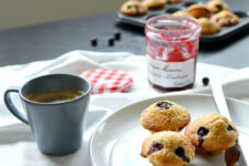 cropped image of a beautifully arranged table setting with a white plate holding three delectable brown butter and blueberry mini muffins, accompanied by a fork, and a steaming cup of coffee