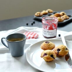 cropped image of a beautifully arranged table setting with a white plate holding three delectable brown butter and blueberry mini muffins, accompanied by a fork, and a steaming cup of coffee