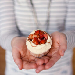 cropped image of a woman holding a single cupcake with fresh cream, strawberries, and balsamic vinegar glaze in both hands