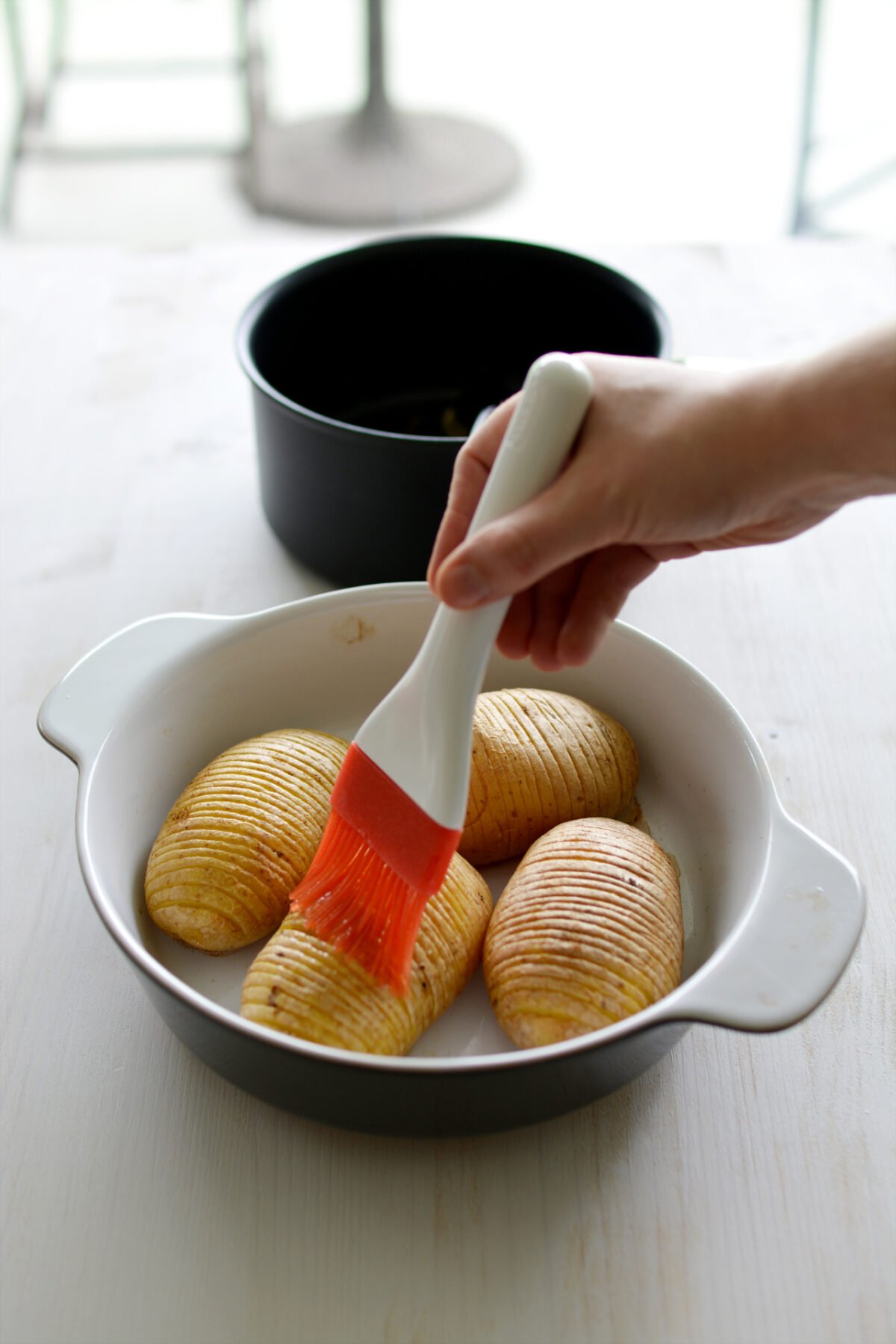 Sliced potatoes (Hasselback style) in a casserole dish with a woman's hand brushing on butter with a silicone brush.