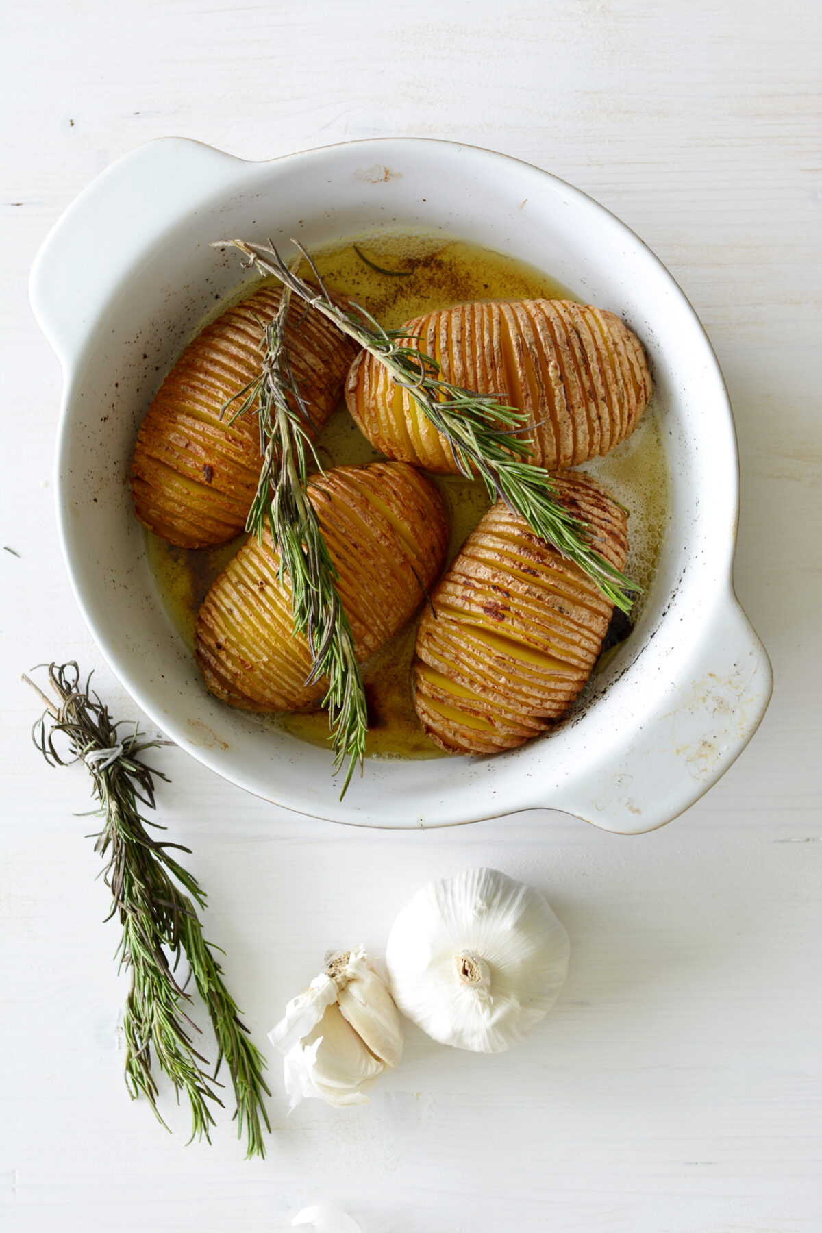 Hasselback Potatoes in a casserole dish on a white table with garlic and rosemary.