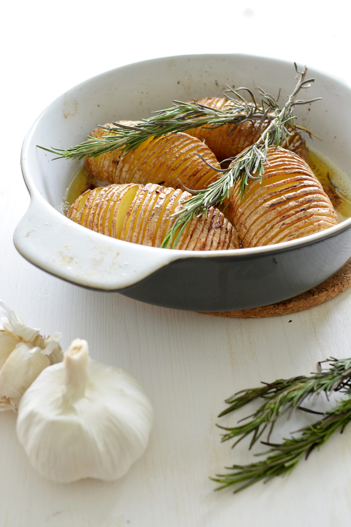 Hasselback Potatoes in a casserole dish on a white table with garlic and rosemary.