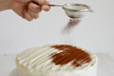 cropped image of a hand using a strainer to sprinkle cocoa powder onto a chocolate cake covered with cream cheese frosting