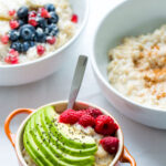 a cropped image of a bowl containing an oatmeal topped with fresh slices of avocado and raspberries