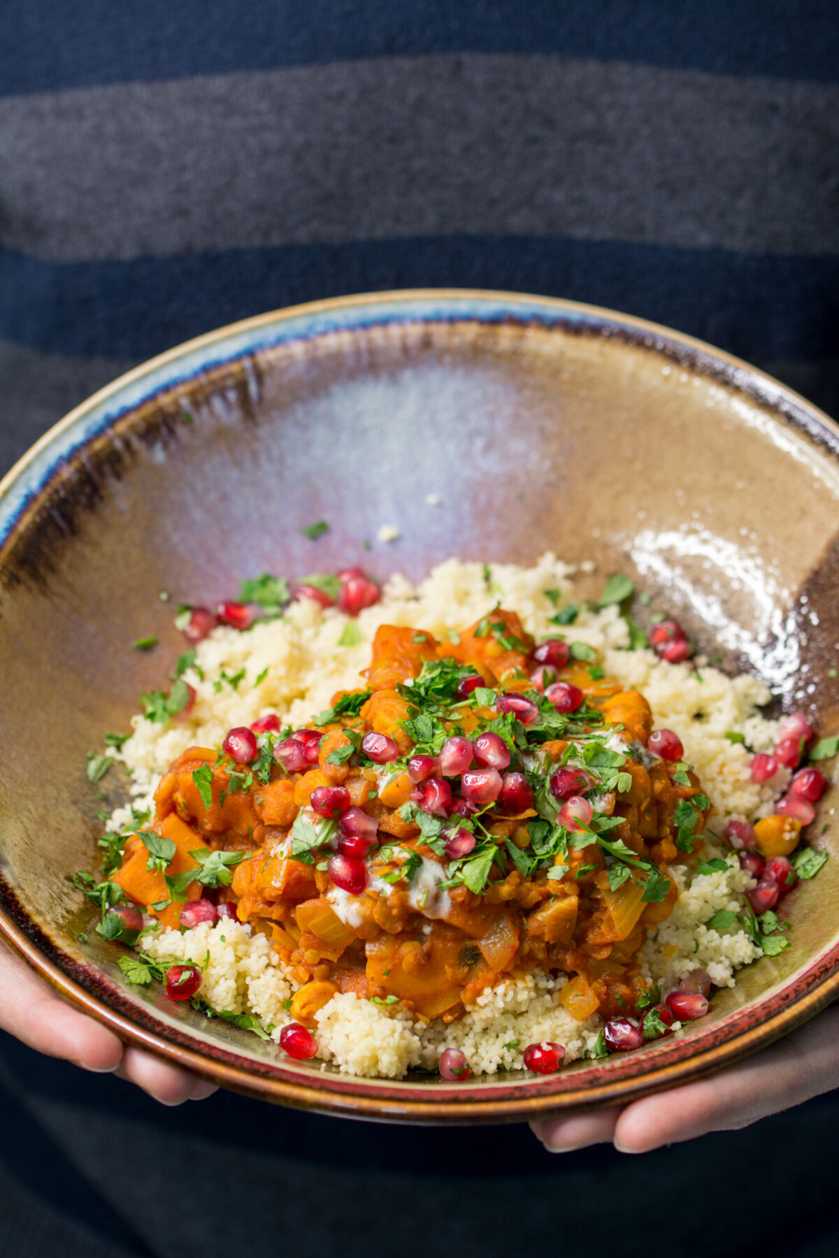 Sweet Potato and Chickpea Moroccan Style Stew in a tan glazed dish topped with pomegranate seeds being held out in a woman's hands.