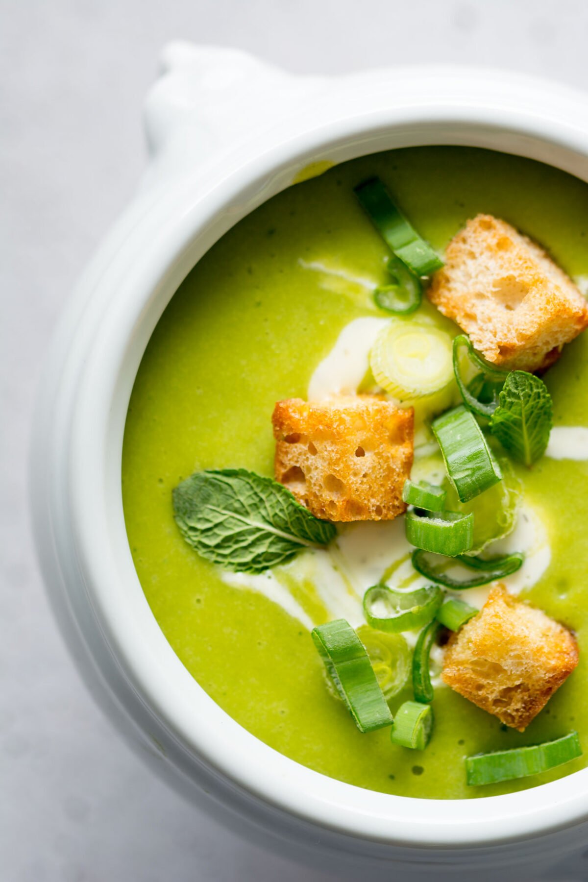 Closeup of a white bowl of mint and pea soup topped with croutons, green onions, fresh mint, and coconut cream on a white background.
