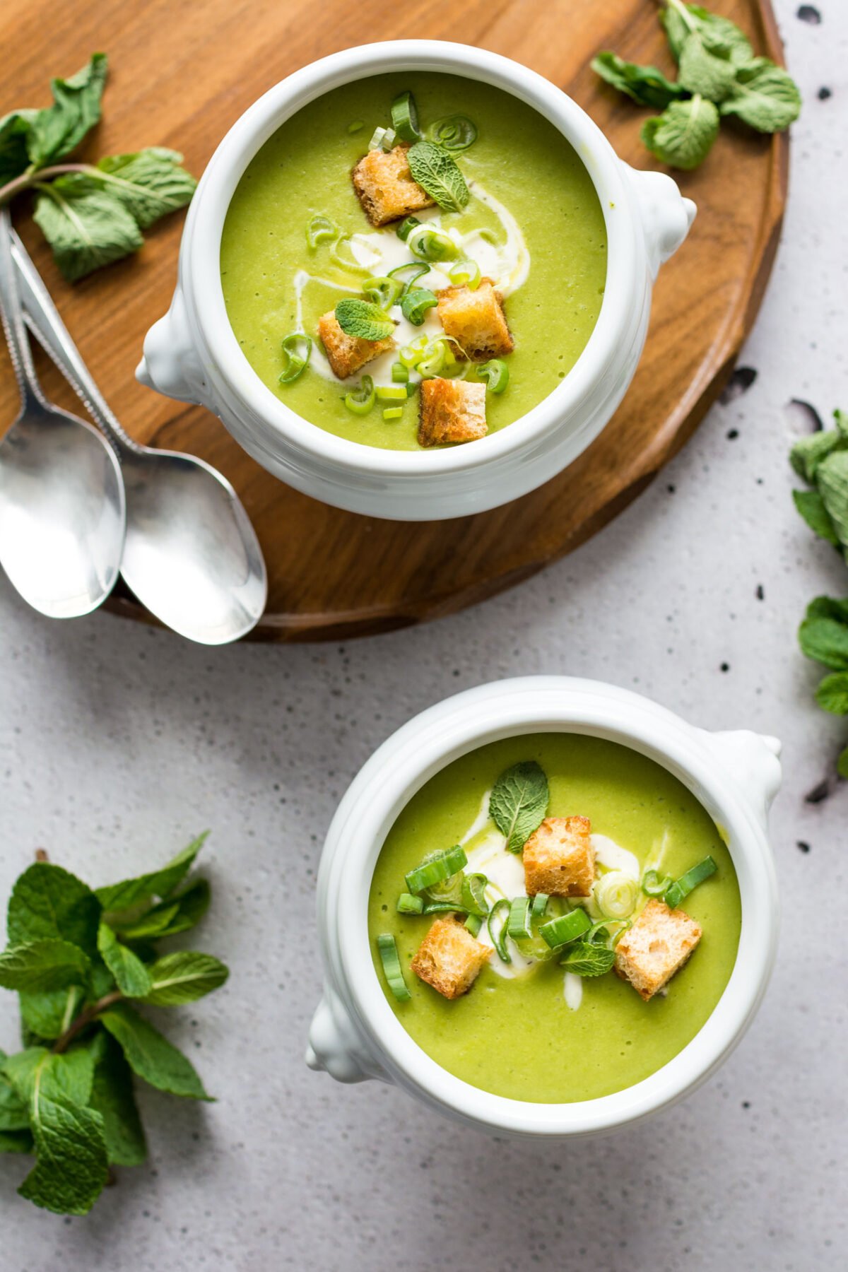 Two white bowls of pea and mint soup on a grey background surrounded with fresh mint and spoons.