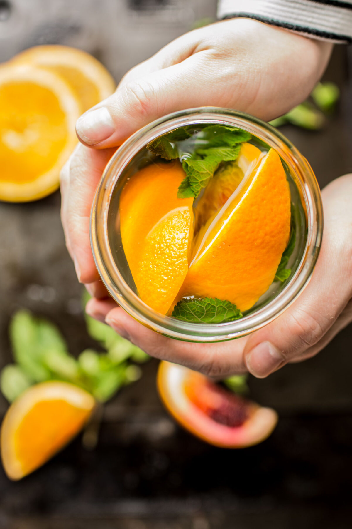 A woman's hands holding a glass jar filled with orange slices and fresh mint sprigs.