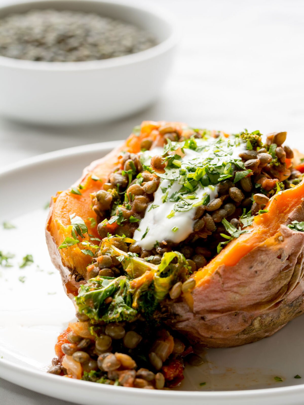 Sweet Potato stuffed with lentils, kale and sun dried tomatoes on a white plate with more lentils in a bowl in the background.