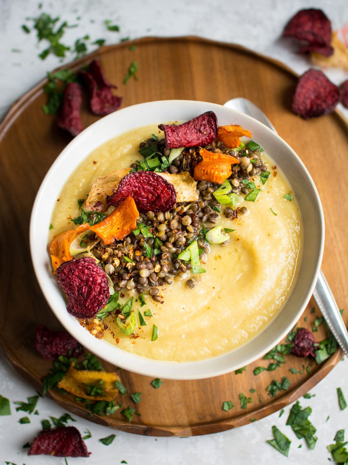 top-down image of a bowl full of delicious, sweet parsnip and apple soup, loaded with lentils and roasted vegetable crisps topped with black lentils, spring onion, parsley, chili flakes, and root vegetable chips 
