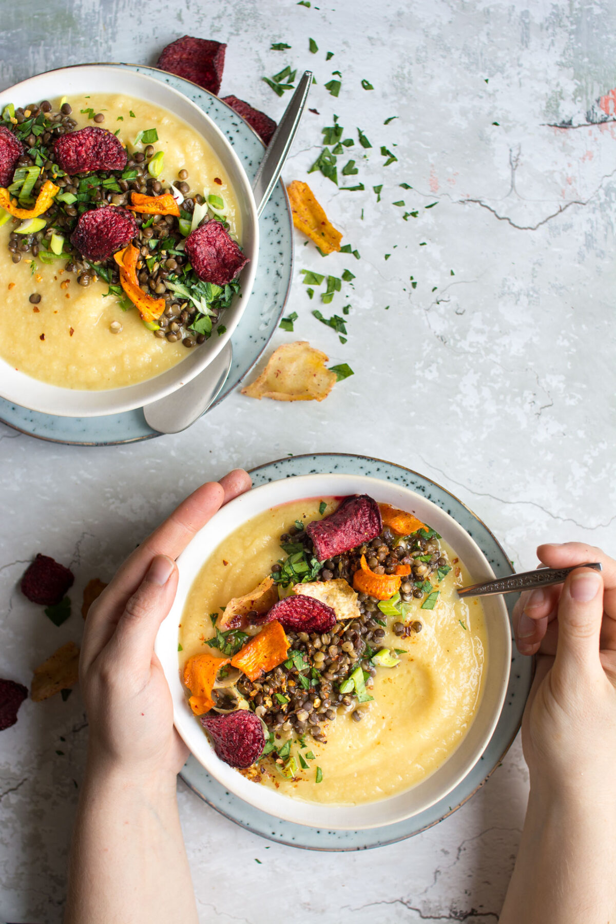 top-down image of a hands holding a bowl full of delicious, sweet parsnip and apple soup, loaded with lentils and roasted vegetable crisps topped with black lentils, spring onion, parsley, chili flakes, and root vegetable chips 
