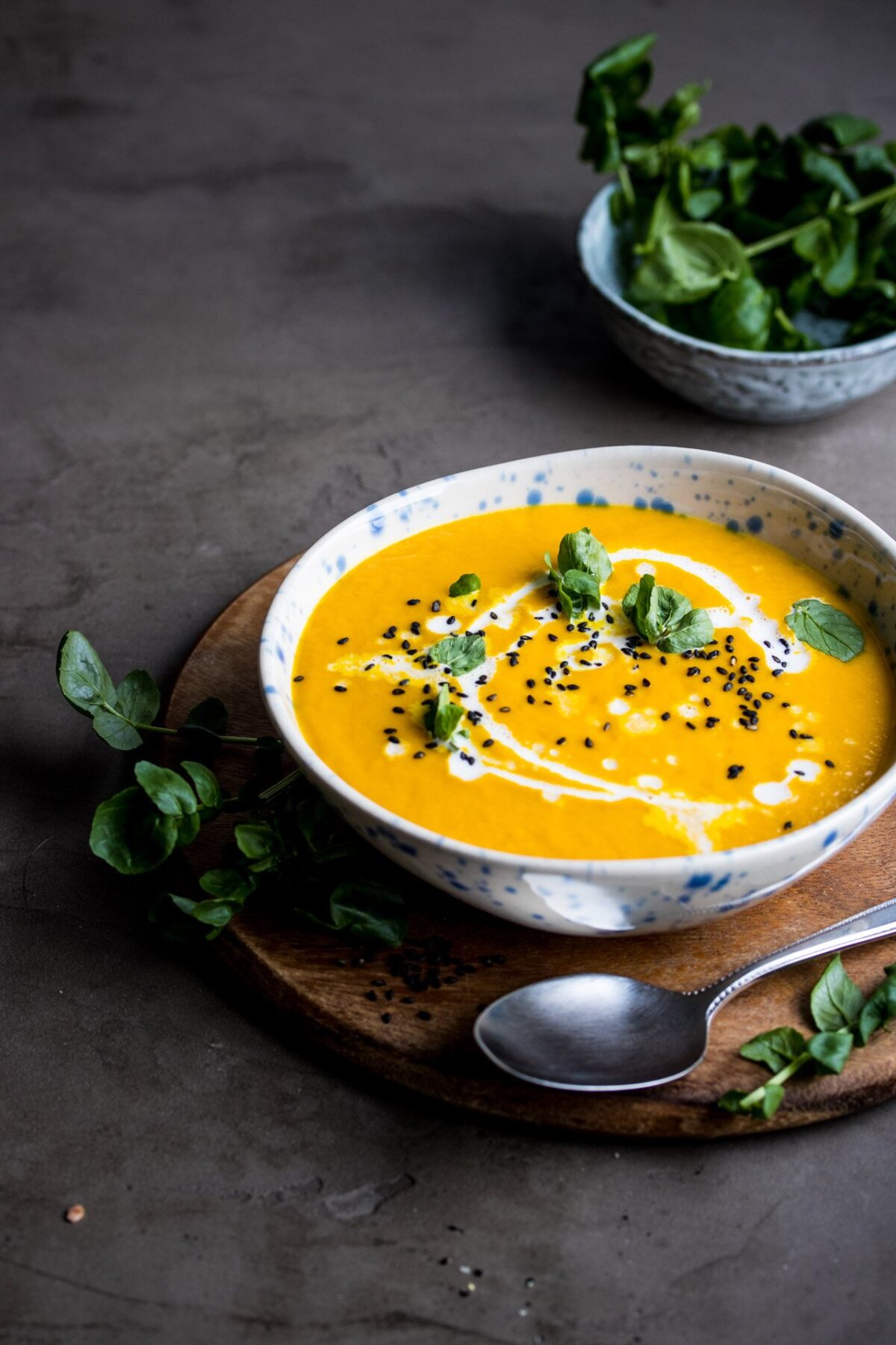 A bowl of turmeric soup with coconut milk drizzled in it. The soup is on a wooden platter with a spoon beside it and some herbs.