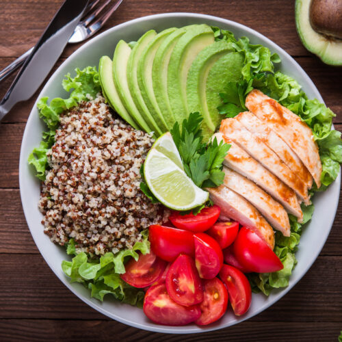 Healthy salad plate with quinoa, tomatoes, chicken, avocado, lime and mixed greens (lettuce, parsley) on wooden background.
