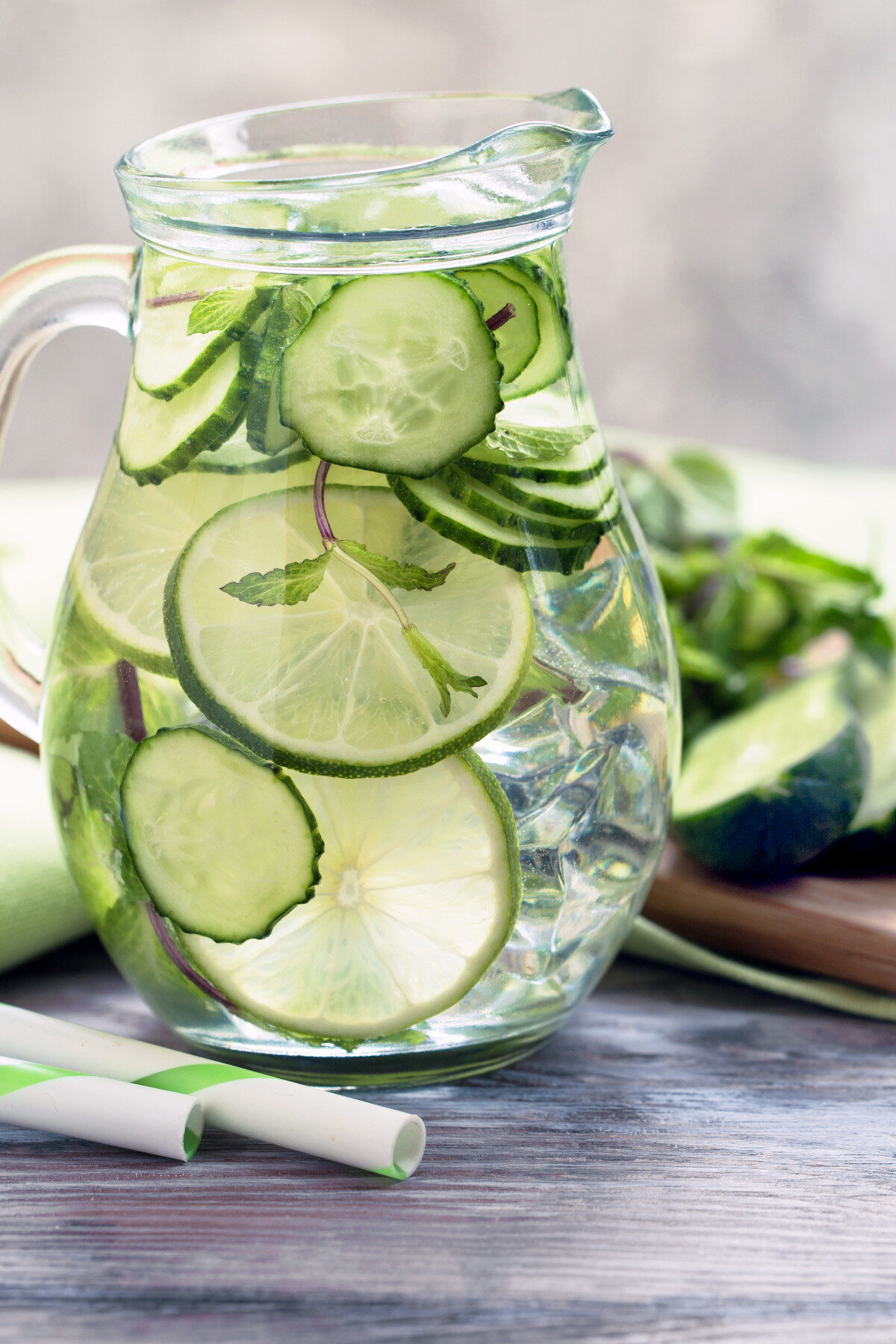 Vertical image of a glass pitcher filled with cucumber water, flavored with lime and mint.