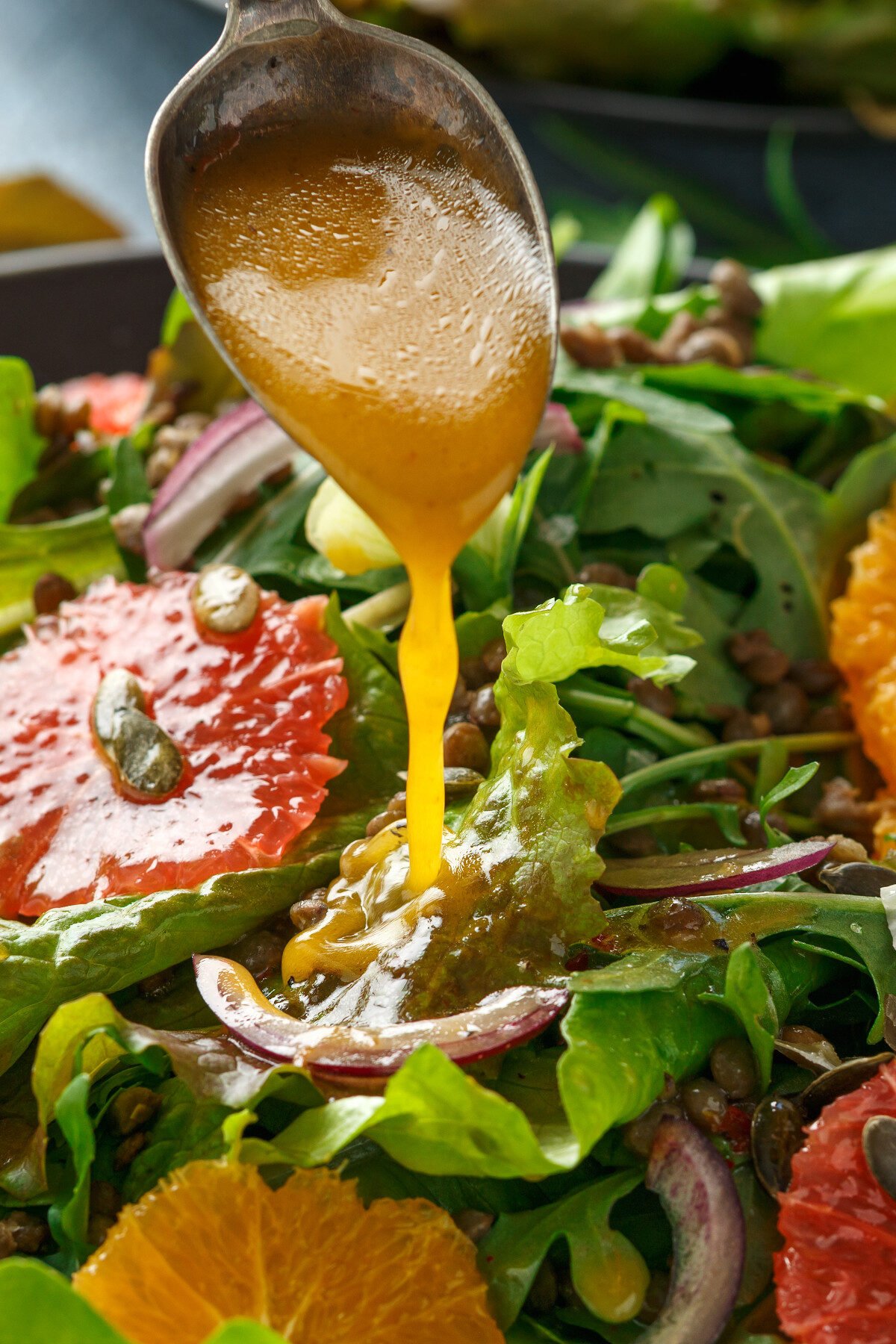 Vertical image of vinaigrette dressing pouring from a spoon onto a fresh fruit salad with mixed greens.