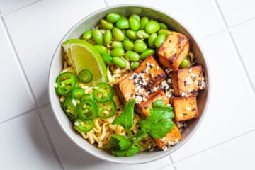 High protein vegetables in a vegan noodle ramen soup with marinated tofu, edamame beans and hot peppers in a gray bowl on a white tile background, top view.