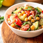 Mediterranean chickpea salad with tomatoes, cucumbers, and onions in a white bowl on a wooden cutting board.