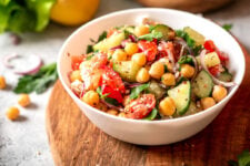 Mediterranean chickpea salad with tomatoes, cucumbers, and onions in a white bowl on a wooden cutting board.