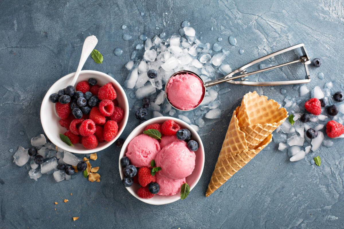 Berry ice cream in a white bowl with fresh blueberries and raspberries in another bowl, along with an ice cream scoop full of berry ice cream and a couple of waffle cones.