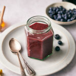 Blueberry Vinaigrette in a glass container on a white plate with blueberries and honey in the background.