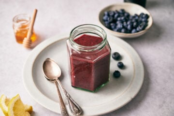 Blueberry Vinaigrette in a glass container on a white plate with blueberries and honey in the background.