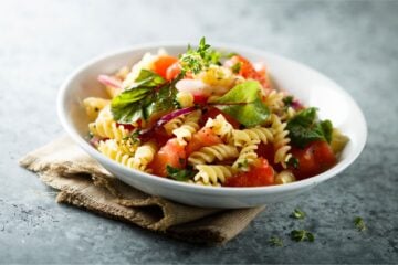 Pasta salad with fresh tomatoes and greens in a white bowl on a burlap napkin on a blue table.