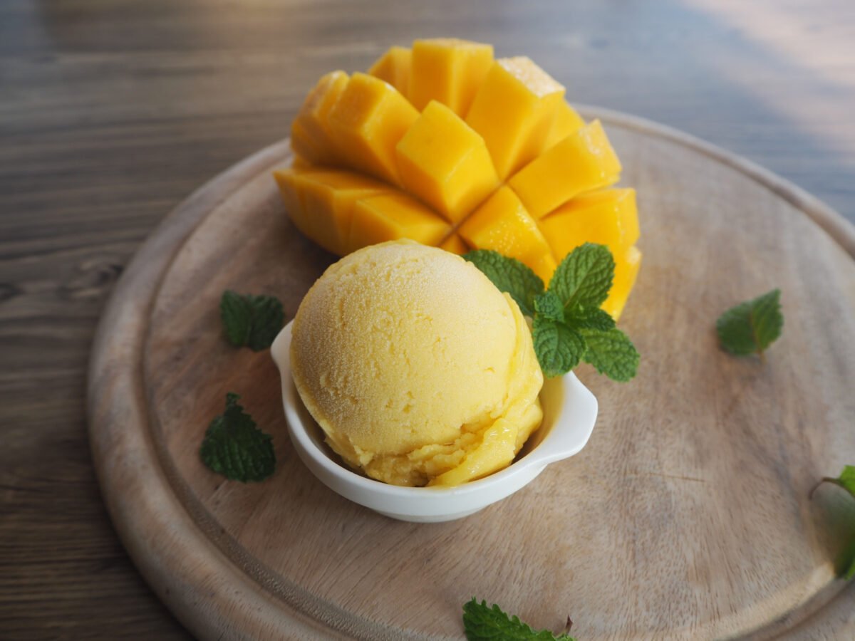 Mango ice cream in a white bowl garnished with mint and with mango slices in the background.