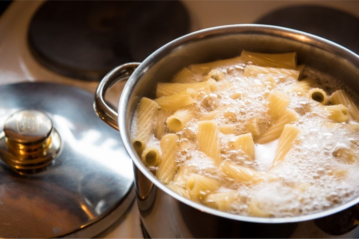 Large saucepan full of boiling water and pasta sitting on a stovetop with the saucepan lid beside it.