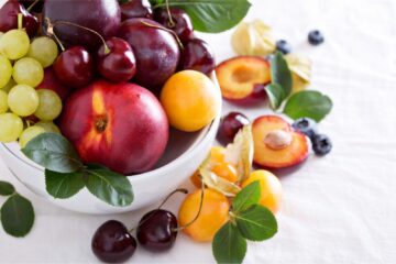 Peaches, plums, green grapes, cherries, apricots in a white bowl on a white cloth background.