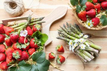 A basket of fresh-picked strawberries and a bundle of asparagus tied with twine on a wooden surface.