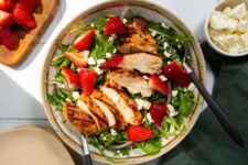 Fresh green salad topped with feta cheese, strawberries, and grilled chicken on a tan and white plate with a white background and a green napkin.