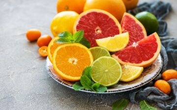 Cut lemons, grapefruit, limes, and oranges on a ceramic plate with more citrus in the background plus a blue cloth napkin on a slate colored table.