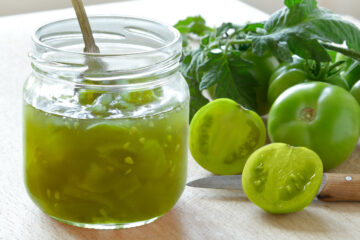 Green tomato chutney in a glass jar with a spoon and green tomatoes in the background.