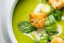 Pea and Mint soup closeup in a white bowl topped with croutons, green onions, and fresh mint leaves.