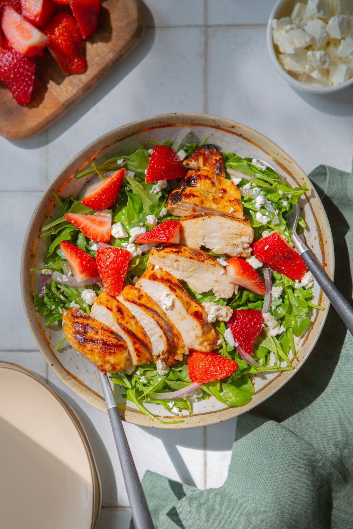 Salad topped with grilled chicken and fresh strawberries on a white tile background with strawberries and feta cheese at the top of the image.