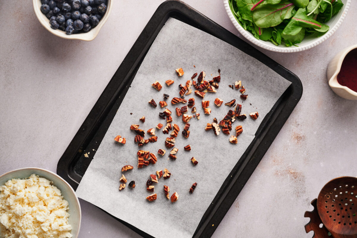 Chopped pecans on a baking sheet with parchment paper with ingredients for a Summer Blueberry Salad.