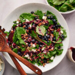 Top flat-lay view of summer blueberry salad with blueberries, feta, and toasted pecans in a white bowl with wooden utensils.