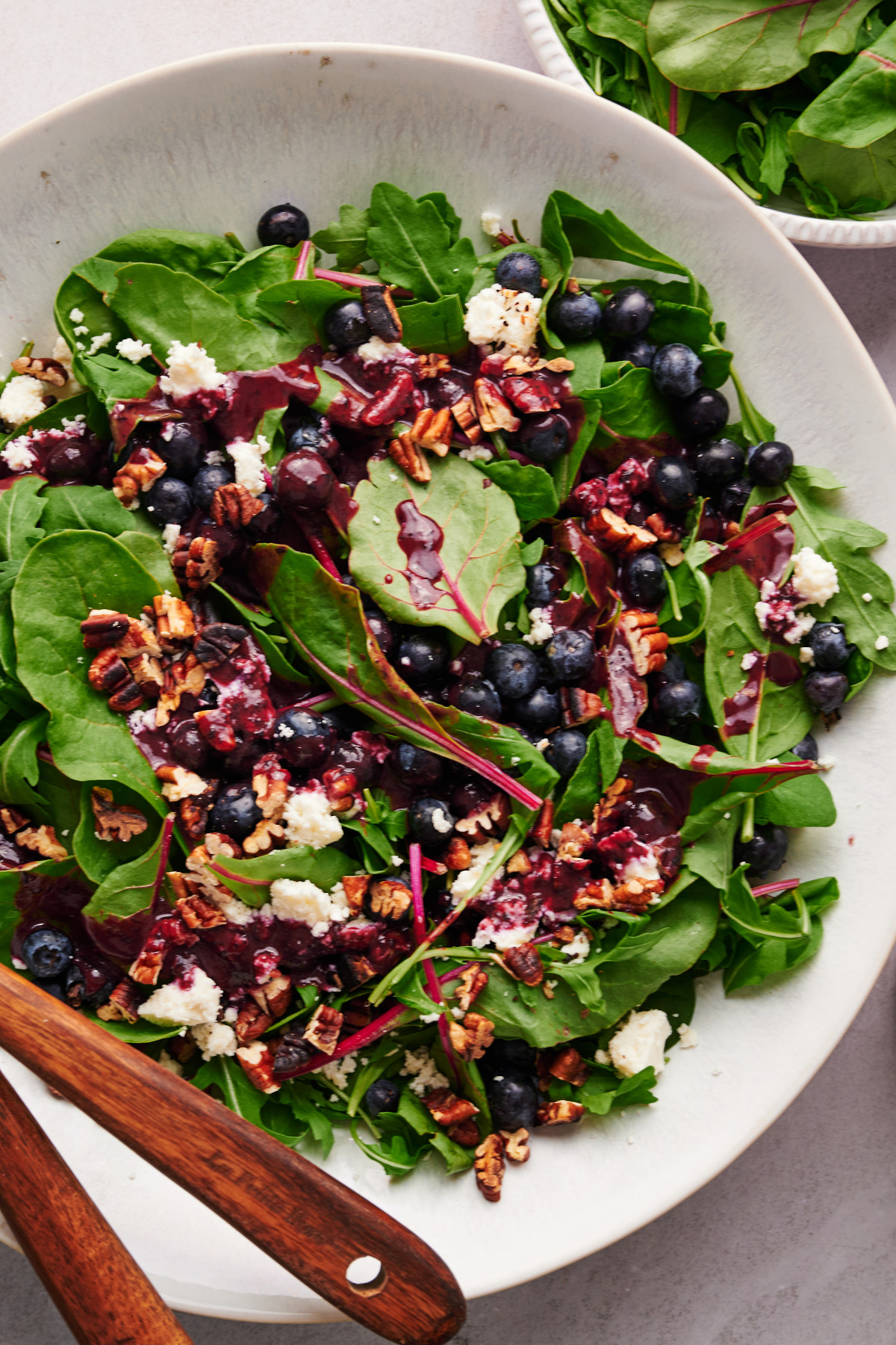 Vertical flat-lay view of summer blueberry salad with blueberries, feta, and toasted pecans in a white bowl with wooden utensils.