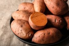 Whole sweet potatoes on a black plate sitting on a burlap tablecloth.