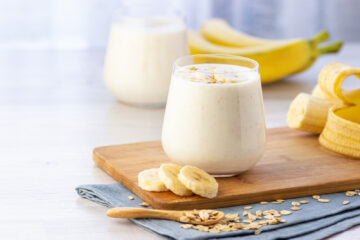 Banana smoothie in a glass on a wooden cutting board with sliced bananas in the background and foreground.