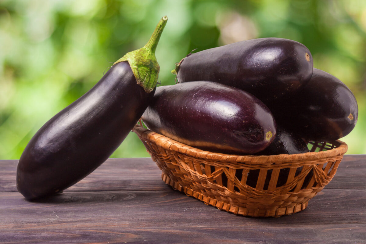 Eggplant in a wicker basket on wooden table with blurred green background.