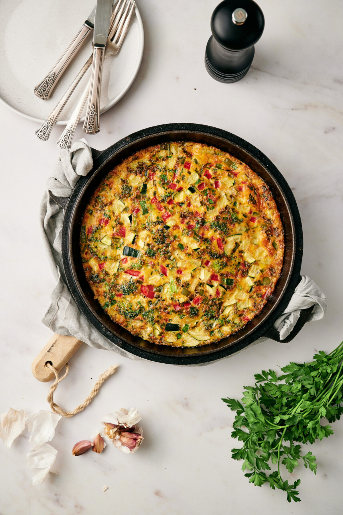 Vertical view of a fully cooked vegetable frittata in a cast iron skillet on a marble countertop with fresh herbs.