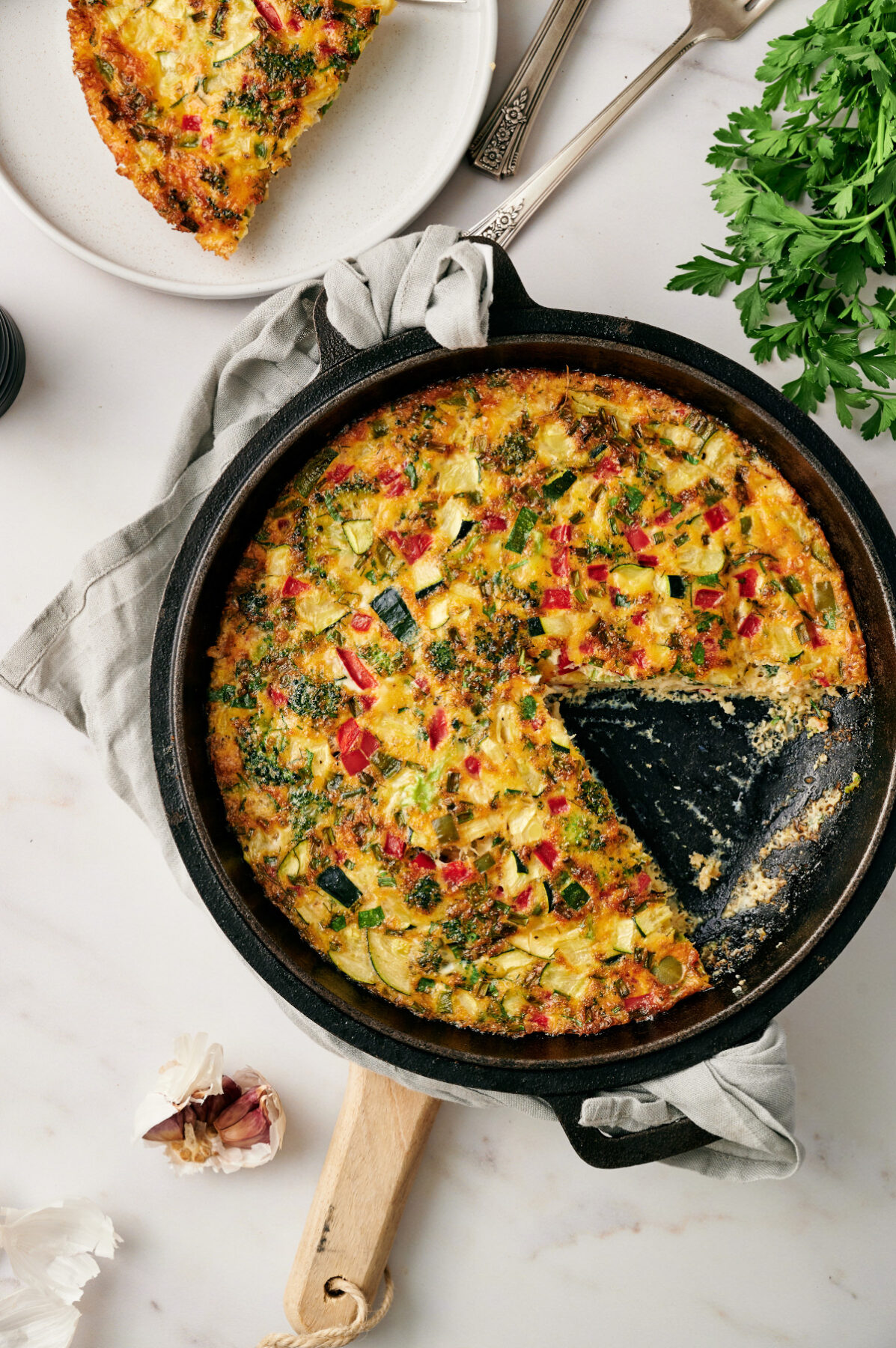 Vertical view of a fully cooked vegetable frittata in a cast iron skillet with one slice removed on a marble countertop with fresh herbs.