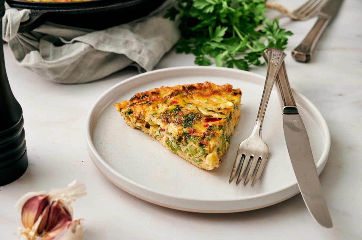A slice of fresh vegetable frittata on a white plate with a fork and knife, with fresh herbs in the background.