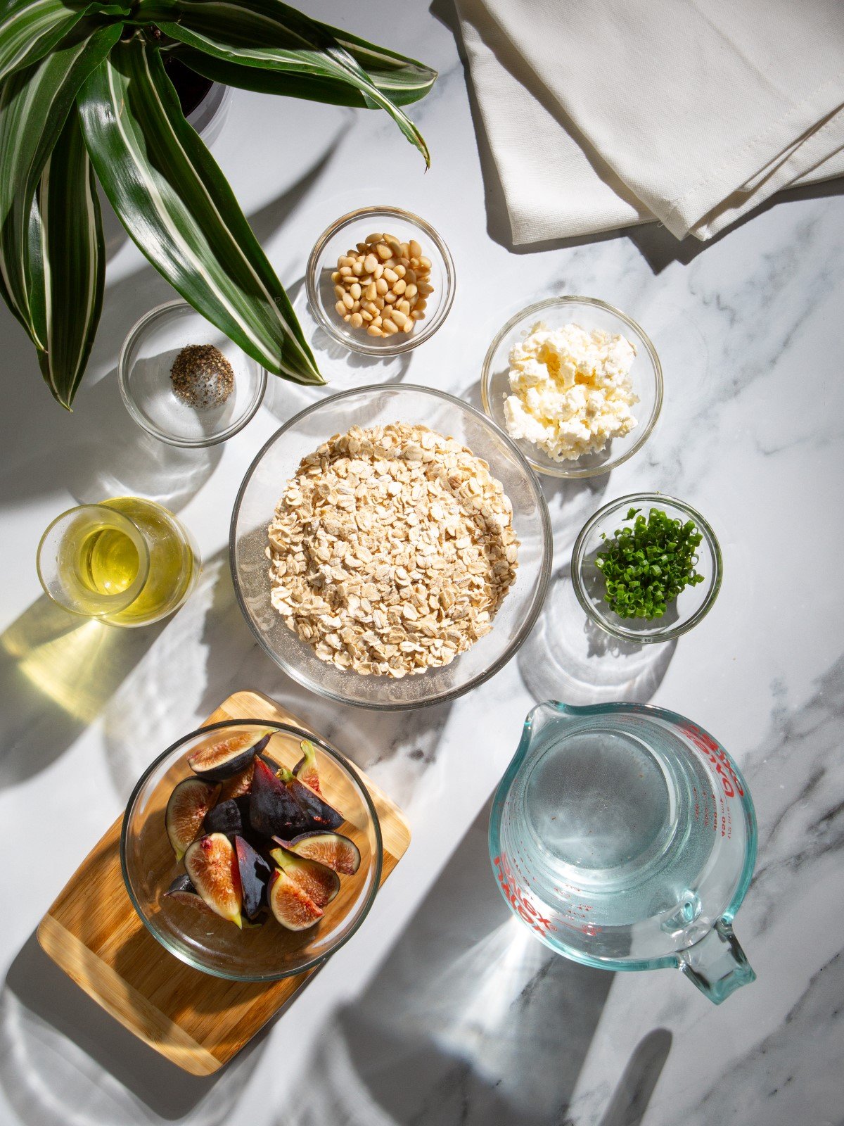 Glass bowls filled with oatmeal, figs, feta, water, and other ingredients with a plant and napkins.