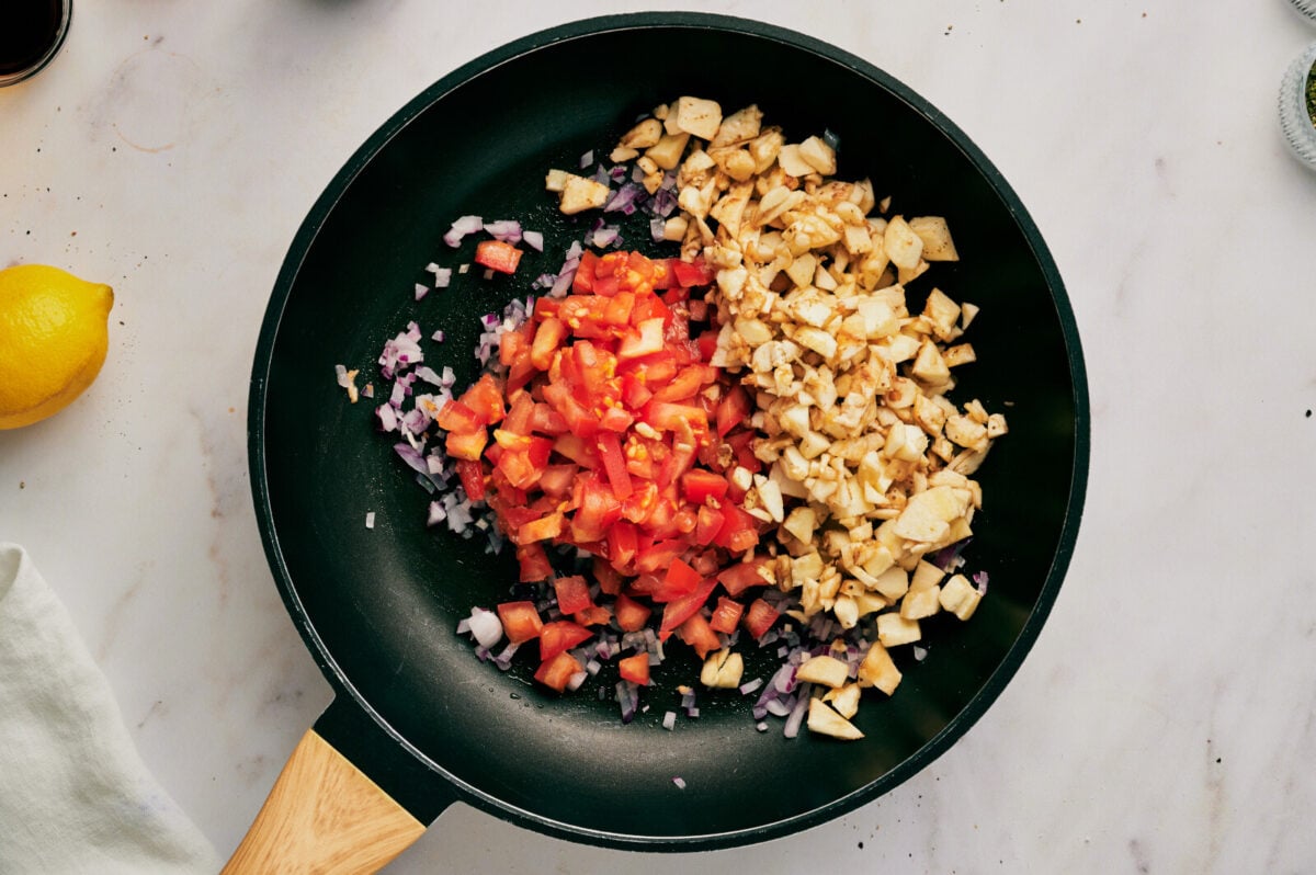 Eggplant, tomato and onion being sautéed in a black skillet.