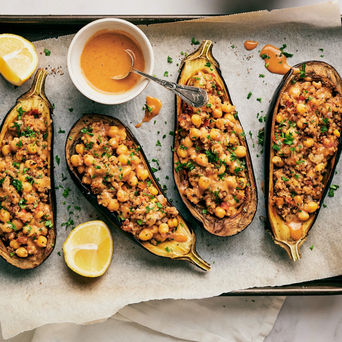 Eggplants stuffed with chickpea and quinoa filling on a baking sheet with parchment paper and a ramekin with sauce and a spoon.