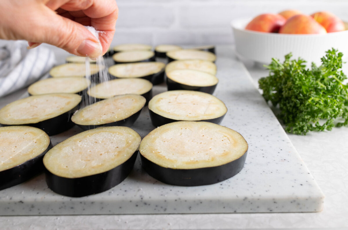 A woman's hand sprinkling salt on slices of eggplant on a cutting board.