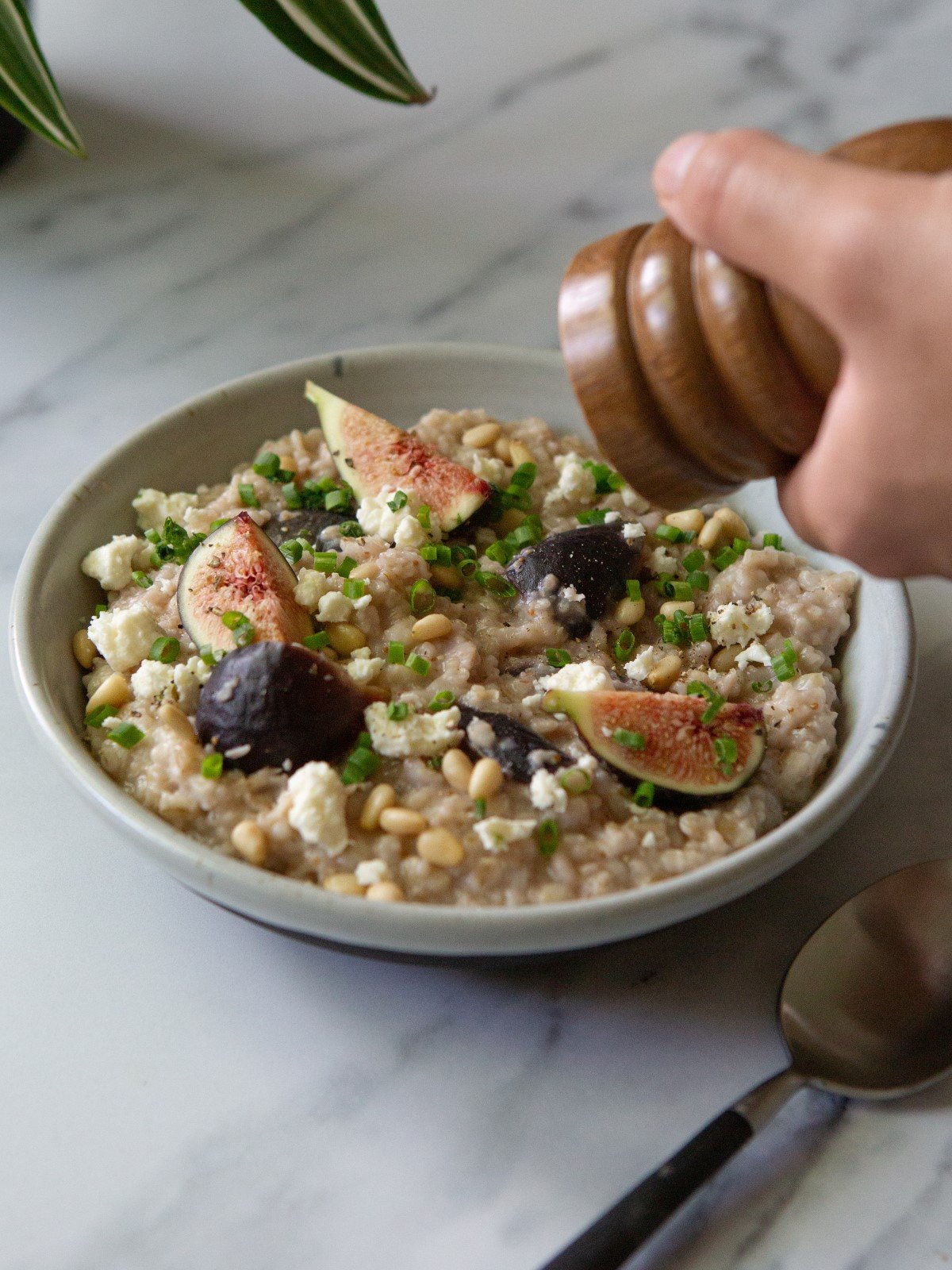 A white bowl filled with savory oatmeal with figs and feta and a spoon. A hand is holding a black pepper grinder.